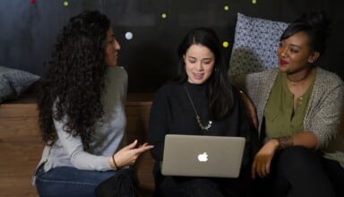 three women sitting on a bench looking at a laptop