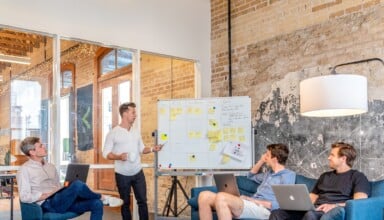 three men sitting while using laptops and watching man beside whiteboard