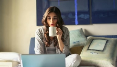 A woman enjoying coffee while working from home in a cozy bedroom setting with a laptop.